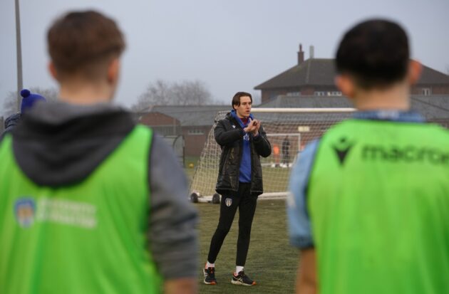 Two boys with their back to the camera on a football field being taught by a coach
