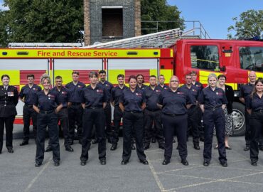 A group of 16 firefighters and control centre officers are standing in a row in front of a fire engine. Deputy PFCC Jane Gardner and Deputy Chief Fire Officer Moira Bruin are standing either side of them.
