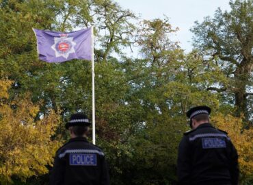Two police officers look towards a blue Essex police flag which is flying at police HQ. They have their backs to the camera and their heads bowed. There are trees in the background and a blue sky.